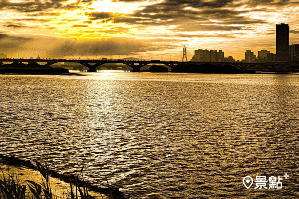傍晚時分，夕陽緩緩自橋邊落下，淡水河面灑染著金黃色彩。